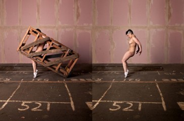 A naked women, is balancing on just her toes. Behind her is a pink tiled wall and the floor is market out with numbers. Is one side of the photo the woman is trapped in a wooden crate.