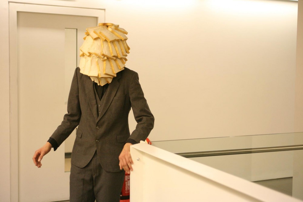 A man in a suit stands at the top of a white staircase, his face is covered in white bread.