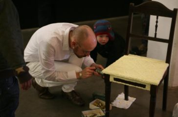 A man dressed in white, is creating a patten on a wooden chair, a toddler is watching him intrigued.