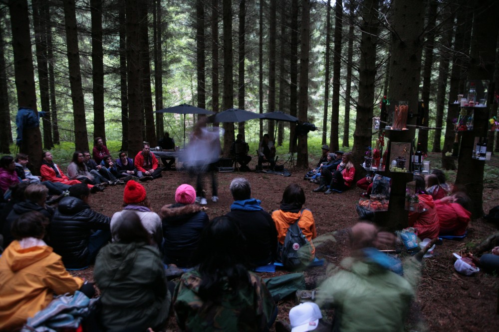 A crowd of people sit in a woodland. A few are holding umbrellas, someone in performing in the centre of the space.