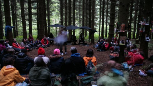 A crowd of people sit in a woodland. A few are holding umbrellas, someone in performing in the centre of the space.