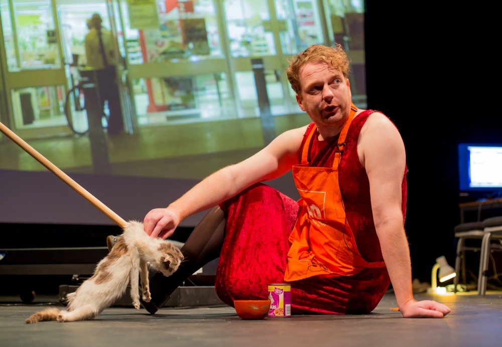 Kim Noble sits on stage wearing an apron, behind him there is a screen playing a video. He is holding a fake cat attached to a wooden poll.