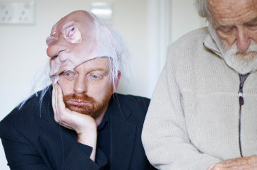 A man sits holding his face in one hand, he is half wearing a mask of an old man on his head. Next to him stands an old man looking down towards the floor.