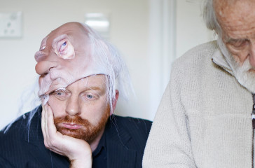 A man sits holding his face in one hand, he is half wearing a mask of an old man on his head. Next to him stands an old man looking down towards the floor.