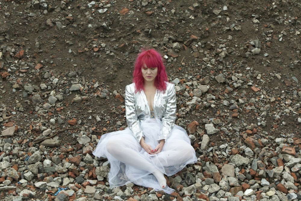 A woman with pink hair is dressed in white and silver, she is sat on the floor surrounded by rubble.