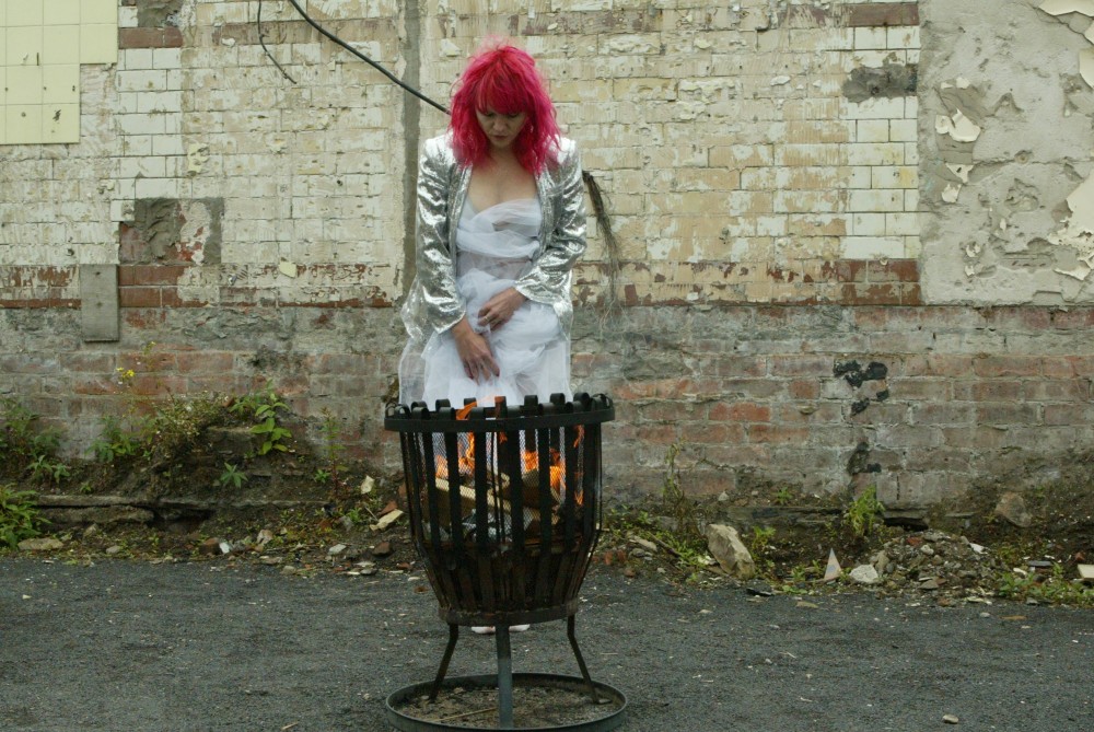 A woman with pink hair is wrapped in material, she is standing in front of a fire pit, looking into it.