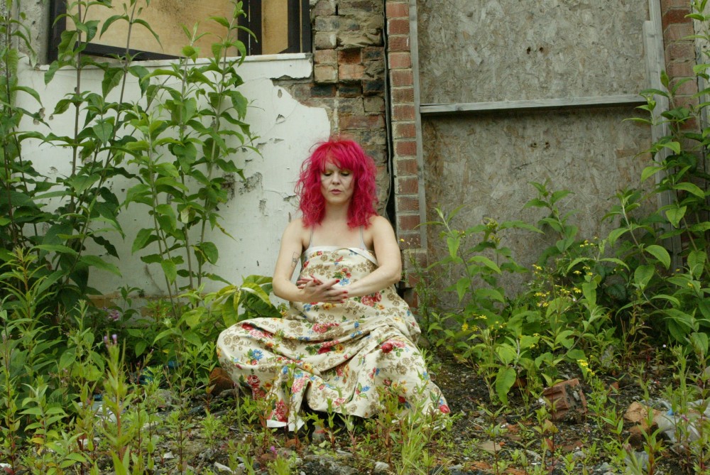 A woman with pink hair, is covered in a piece of flowery pattered material. She is knelt down in a weeded garden.