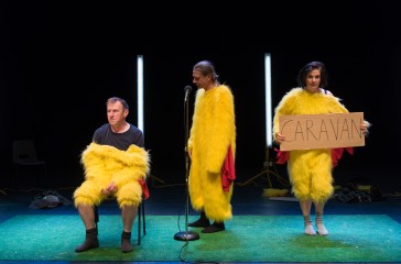 Two men and a woman are dressed in chicken suits are on a stage. One man stands in front of a microphone, one man is sat down and the woman is holding a sign that reads 'Caravan'