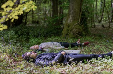 Two people lay in a woodland, they are wearing headphones they lay peacefully surrounded by trees.
