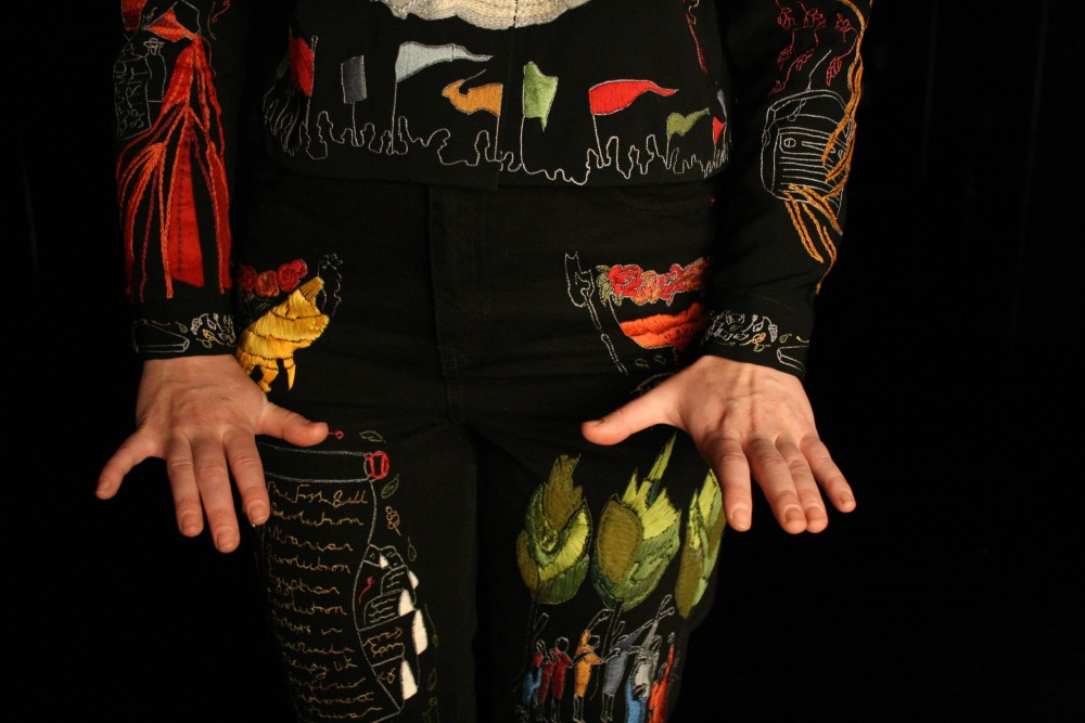 Someone dressed in vibrant colours and pattens, is stood against a black background and has their hands in front of them.