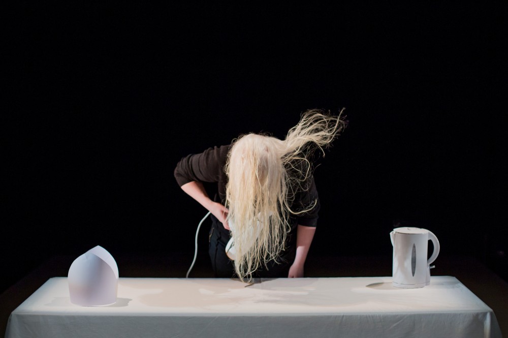 Jo Bannon is drying her hair, in front of a white table. On the table is a white object and a white kettle.