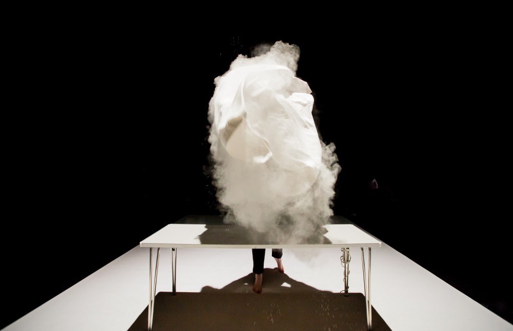 Joe Bannon is in front of a white table on stage. She has just lifted a white cloth into the air, the cloth contains white powder which has been thrown into the air.