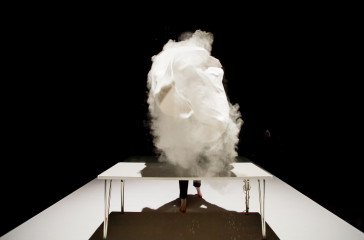 Joe Bannon is in front of a white table on stage. She has just lifted a white cloth into the air, the cloth contains white powder which has been thrown into the air.