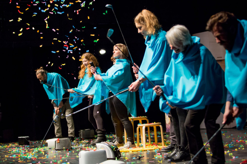 Six people stand on stage dressed in bright blue ponchos, each of them are holding a golf club, they are hitting multi coloured confetti into the air.
