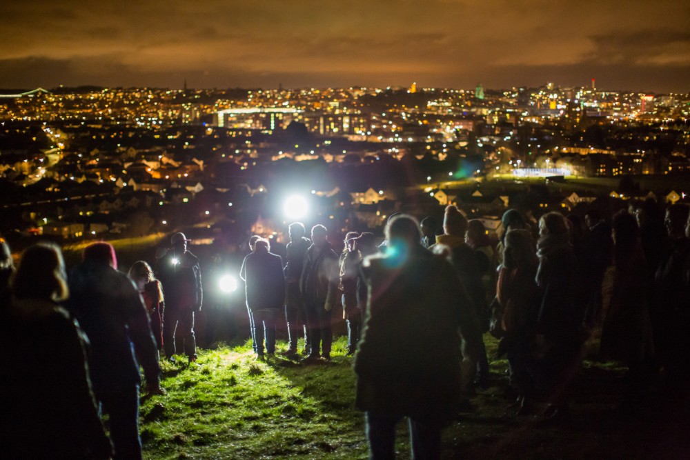 A crowd of people are standing on a hill, their are torches shining up the hill towards the camera. In the background you can see the city lit up.