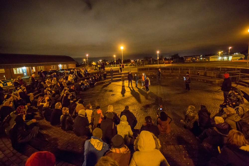 A crowd of people are standing and sitting in a outdoor arena, it is lit by street lights and six Teenagers are stood in the centre.