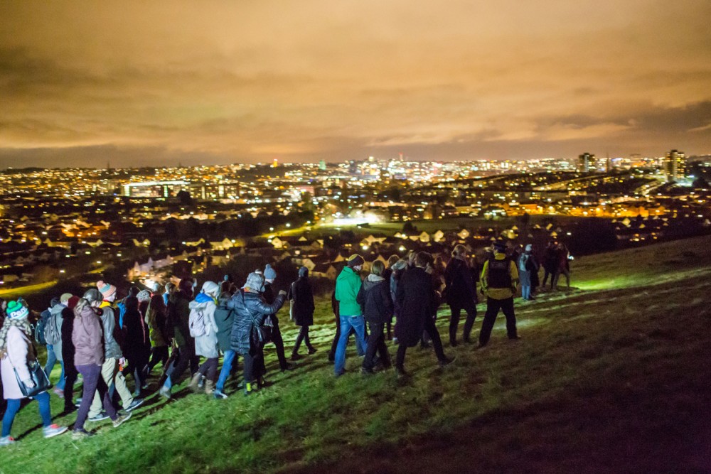 A crowd of people walk across a hill at night, in the background you can see the city lit up.