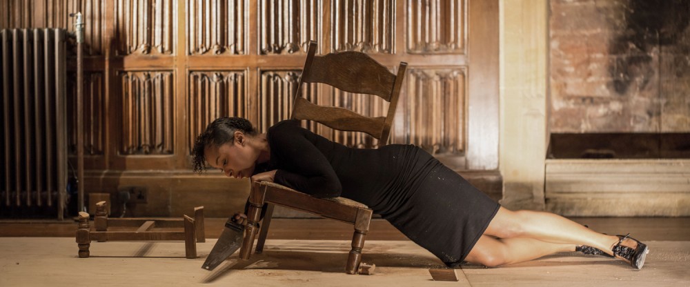 A woman is laid out on the floor, she is partially resting on a wooden broken chair, which is tilted to the side.