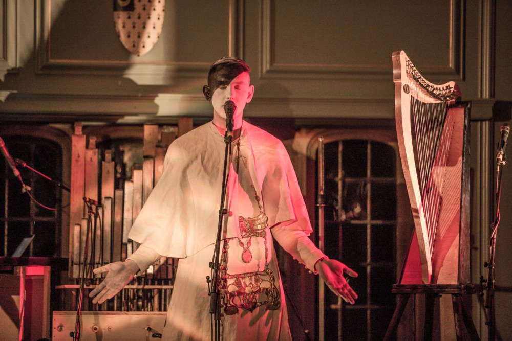 Patrick Wolf stands centre stage, he is painted half black and half white and is wearing a full white costume. He stands in front of a microphone and has his arms held out and his eyes closed. He is surrounded by instruments.