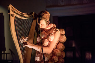 A man wearing a knitted outfit, sits playing the harp.