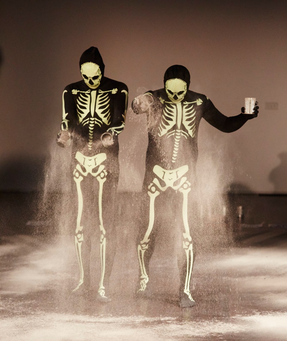Two men are dressed in skeleton skin suits. They are sprinkling white powder onto the floor.
