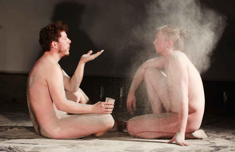 Two naked men are sat on stage, the man on left is blowing white powder into the other mans face.