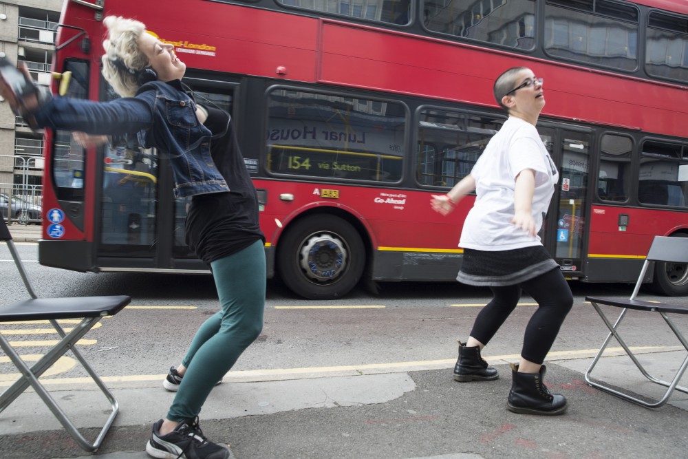 Two women are dancing in the street as a bus goes past them.