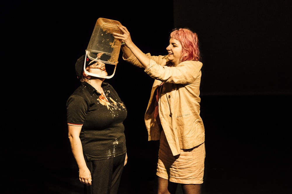A woman with pink hair, is pouring a bucket of baked beans onto Katy Baird on stage.