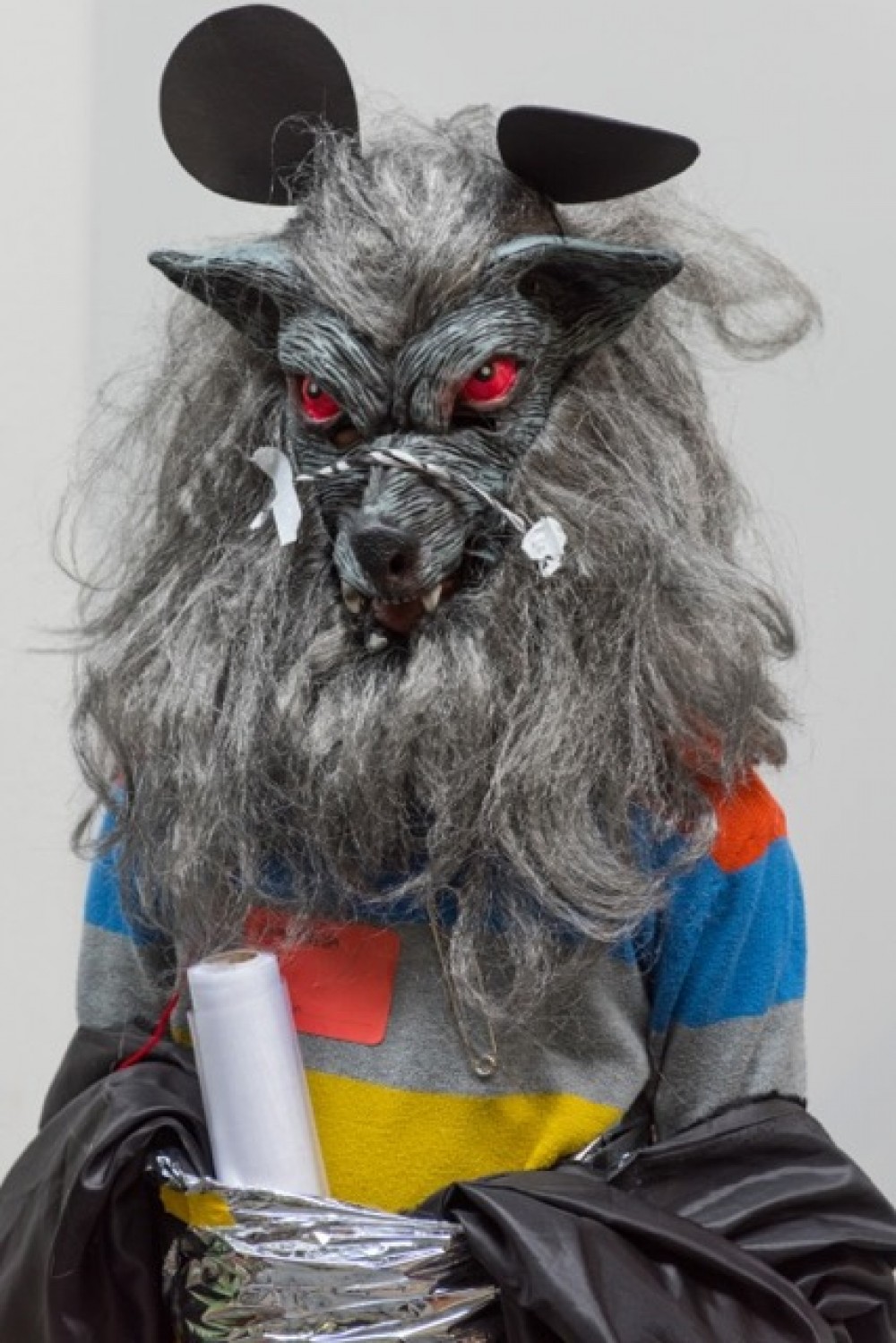Someone wearing a stripped top and a werwolf mask is centred in the photograph. They have a role of plastic tucked into their clothing.