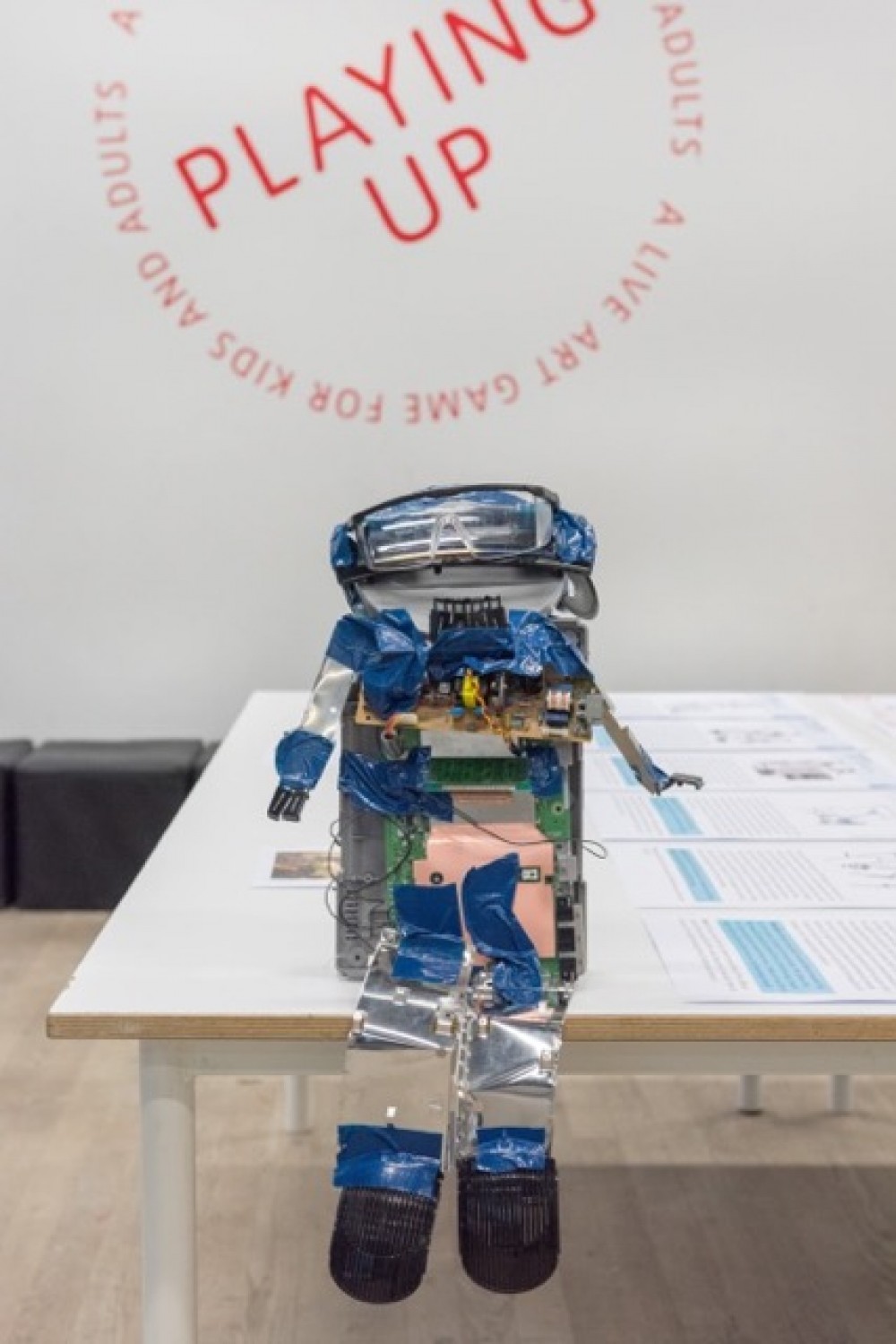 A robot, sits on a table facing the camera. Behind it the logo of LADA Playing Up is posted on the wall.