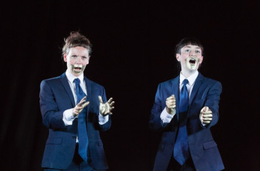Two women dressed in blue suits, have gold painted across their mouths and their hands. They are speaking out towards the audience.