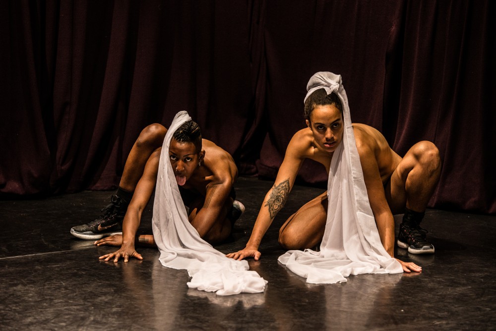 Two women are half dressed and are low on the floor, they have pieces of white material wrapped around their hair and they are strong in their pose.