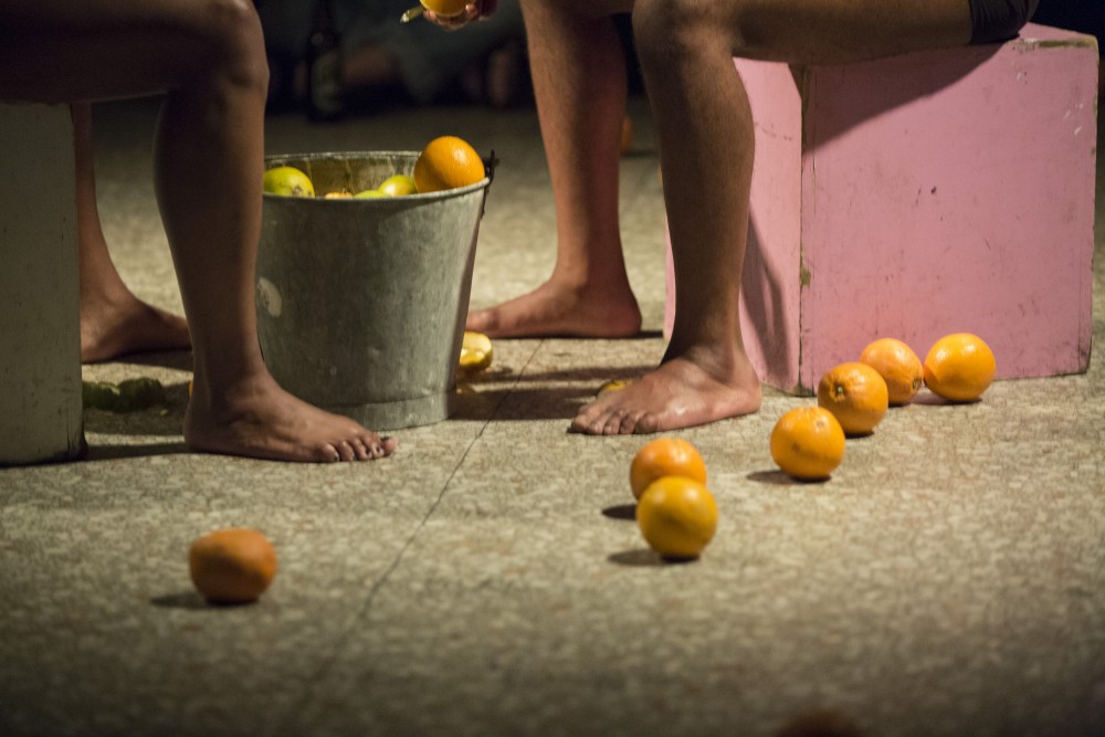 The legs of two people facing each other, with a bucket in front of them, their oranges are on the floor around them.