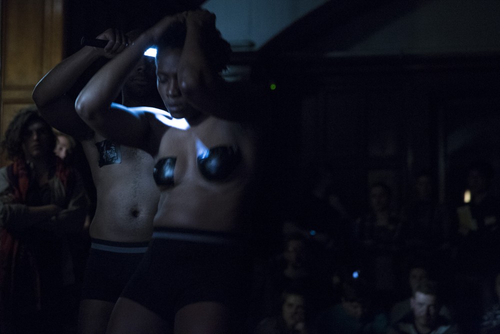 Two people stand in front of an audience, they are half dressed and are holding onto their heads, they have black tape across their chests.