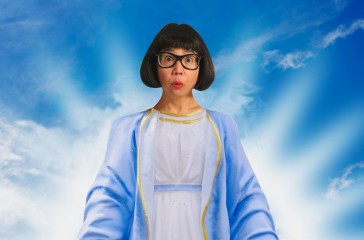 Stacy Makishi is dressed in Jesus style clothing, and is centred in the middle of a blue clouded sky.