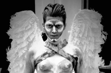 A man covered in white paint, wearing a pair of white wings stares directly into the lens.
