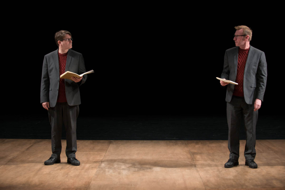 Two men wearing grey suits and red jumpers stand on stage facing each other and holding brown notebooks.