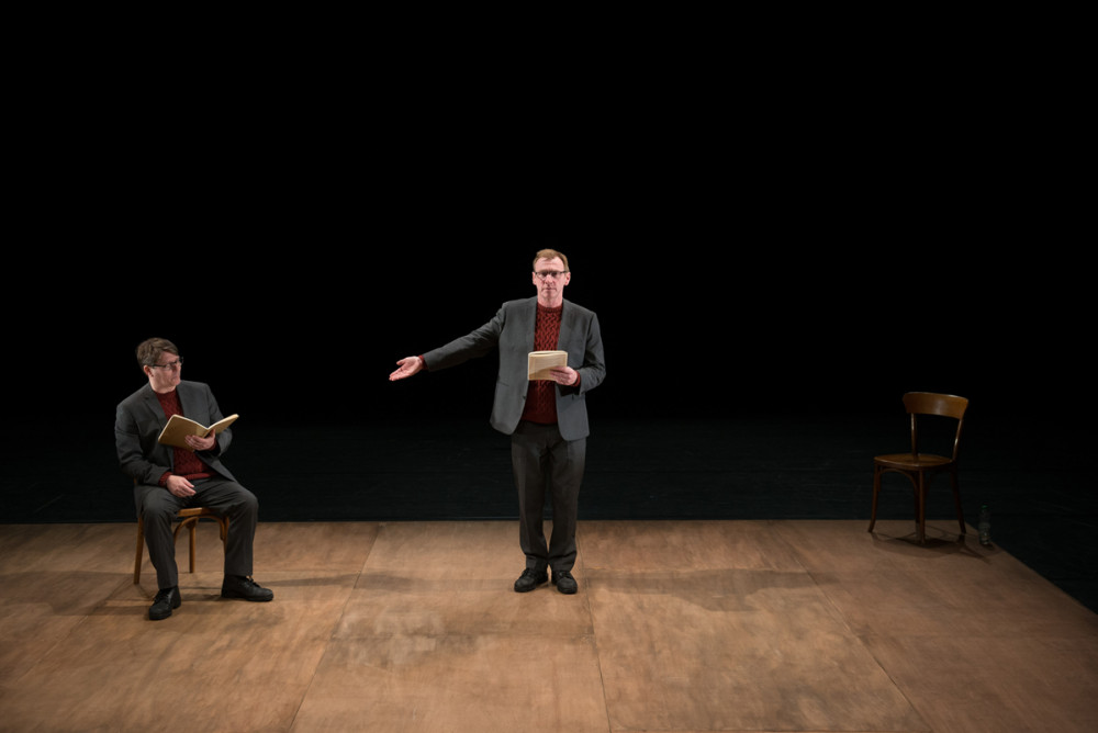 Two men both dressed in grey suits, are wearing red jumpers. One is stood facing the audience, holding his hand out to the other man who is sat on a chair, they are both holding brown books.