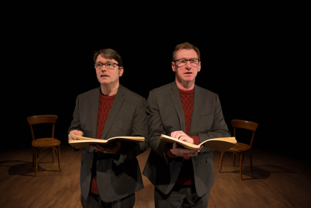 Two men both dressed in grey suits, wearing red jumpers are stood facing the audience holding brown books.