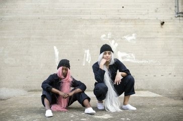 Two girls crouch down wearing overalls and long wigs under beanie hats