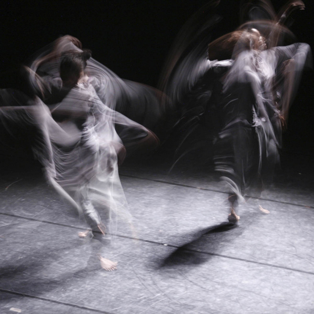 Two figures dressed in grey dance, the image is blurred by the slow shutter release.