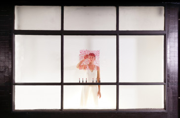 A woman in a white dress stands at a window, she is writing on the window with lipstick.