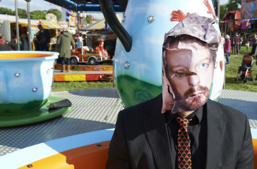 Man sits on fairground ride wearing a mask with an image of a face on it