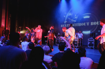 A hip-hop artist speaking into the microphone at the front of the stage whilst others dance.