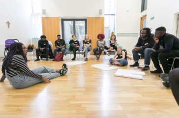 A women sits on the floor speaking to a group who sit in a semi circle around her.