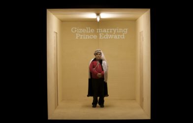 A boy stands in an empty room. He wears a medieval looking headpiece. On the back wall the words 'Gizelle marrying Prince Edward' are projected.