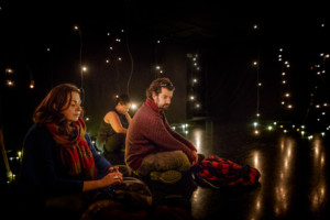 Three people sit cross legged on the floor of a dark room. They are surrounded by hanging lightbulbs. The light reflects on the shiny black floor.