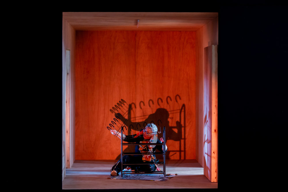 A woman sits on the ground constructing a shoe rack. She is lit from the front with red lighting and cast a dark shadow on the wall behind her.