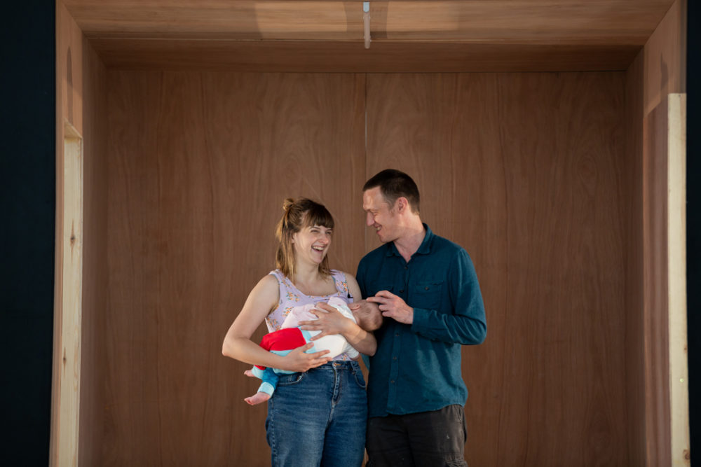 A young woman and a man look at each other laughing. She holds a baby to her chest. He is stroking the babies head.