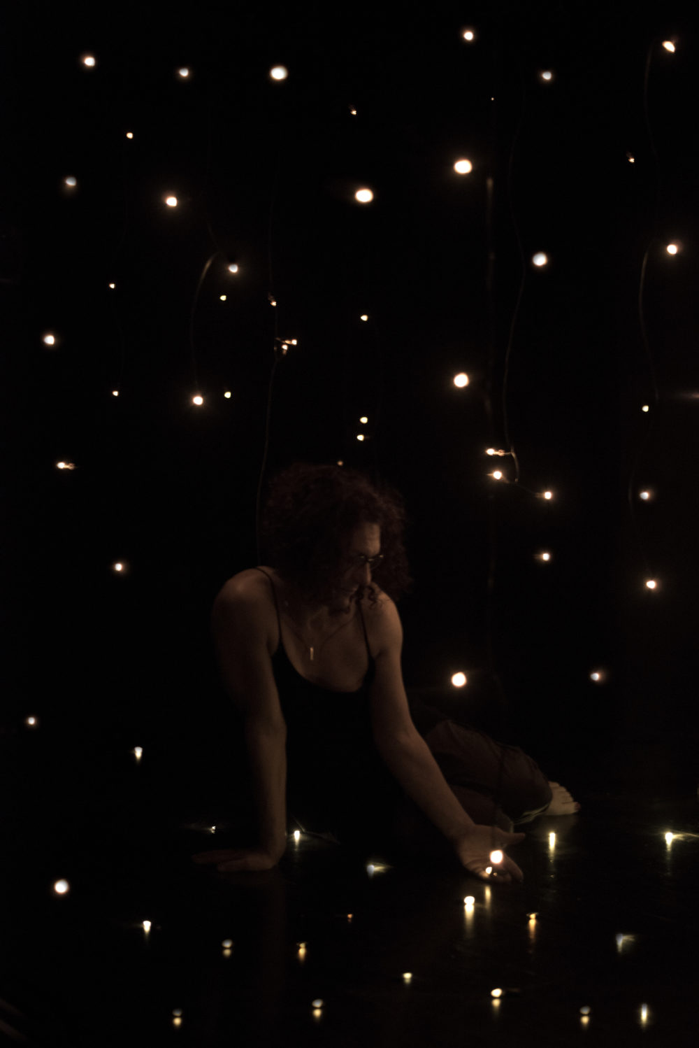 A woman sits in dark room surrounded by tiny lights. She holds one of the lights in an open hand.